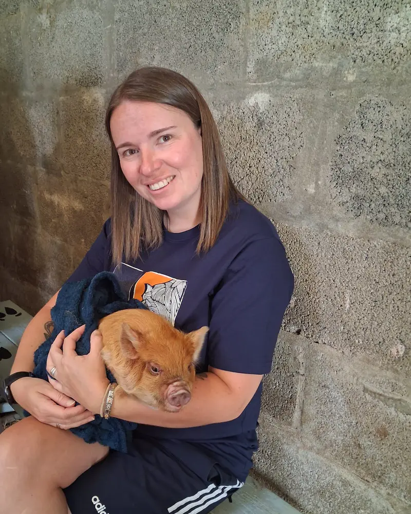 Aimee sitting on a bench, smiling and holding a ginger piglet wrapped in a blue towel